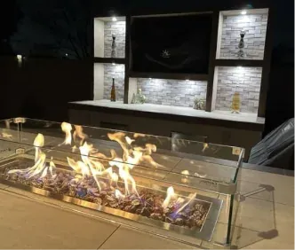 Glass-walled fire pit table burning at night beside a lit stone wall